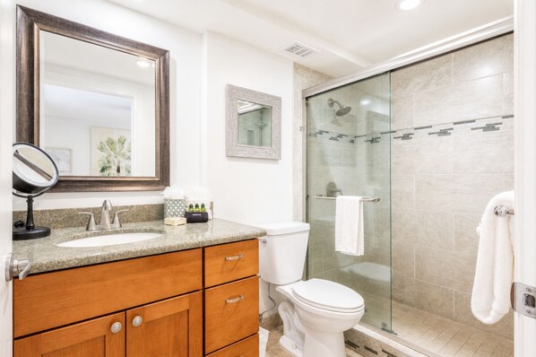 Un baño con encimera de granito, tocador de madera, inodoro blanco, cabina de ducha de vidrio y un espejo grande.