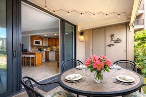 Un espace repas extérieur avec une table ronde prévue pour quatre personnes, entourée de chaises. Une porte vitrée donne accès à une cuisine équipée d’un micro-ondes et de placards. L’espace repas est décoré de guirlandes lumineuses et comprend une petite table sur laquelle est posé un vase de fleurs.