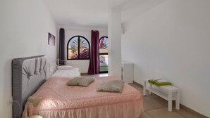 Un dormitorio con cama, mesita de noche, banco, ventana con cortinas y vistas al exterior.