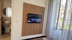 Un dormitorio con cama, televisor montado, baño visible a través de una puerta abierta y ventana con cortinas.