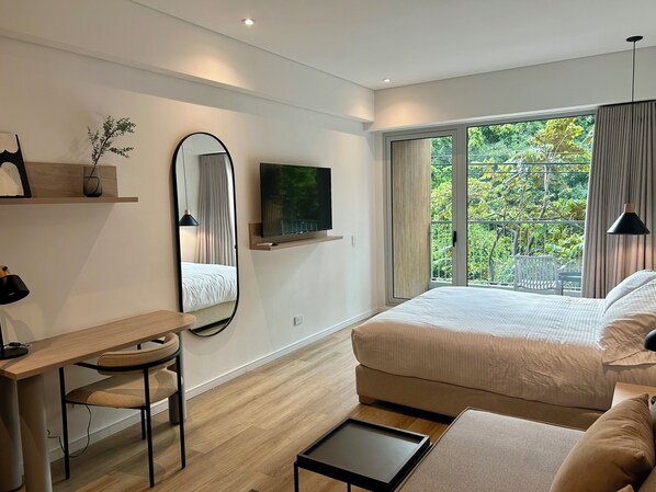Une chambre moderne comprenant un lit, un bureau, une chaise, une télévision et un balcon avec vue sur la verdure.