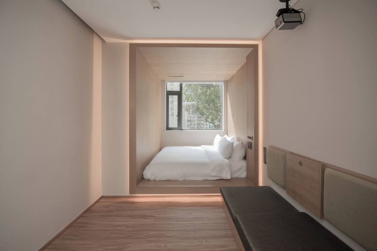 Ein schmales Zimmer mit einem Bett, einer Sitzbank und einem Fenster, das für natürliches Licht sorgt.