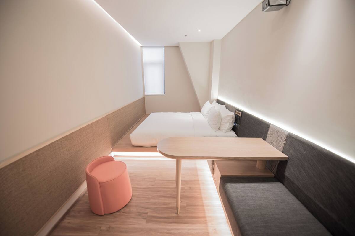 Ein modernes, kompaktes Schlafzimmer mit Bett, einem kleinen Tisch, einer Sitzecke und einem Fenster.