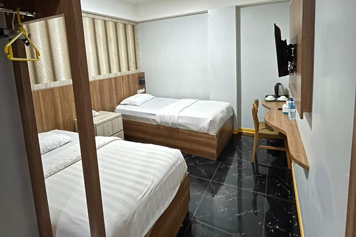 Habitación de hotel con dos camas, un escritorio de madera, una silla, perchero y una balanza pequeña.