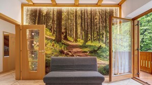 A room with a large window, a grey sofa, and a forest mural.