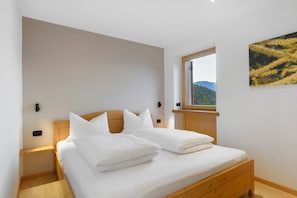 Un dormitorio con cama de madera, ropa de cama blanca, una mesita de noche, una lámpara de pared y una ventana con vistas a zonas verdes.