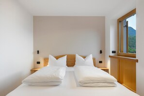 A bedroom with a wooden headboard, two white pillows, a neatly made bed, a nightstand, a wall-mounted light, and a window with a view of a mountain.