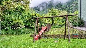 A playground with a slide and swings, surrounded by lush greenery and a mountain backdrop.