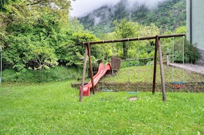 Um parquinho com escorrega e balanços, cercado por vegetação exuberante e uma paisagem montanhosa ao fundo.