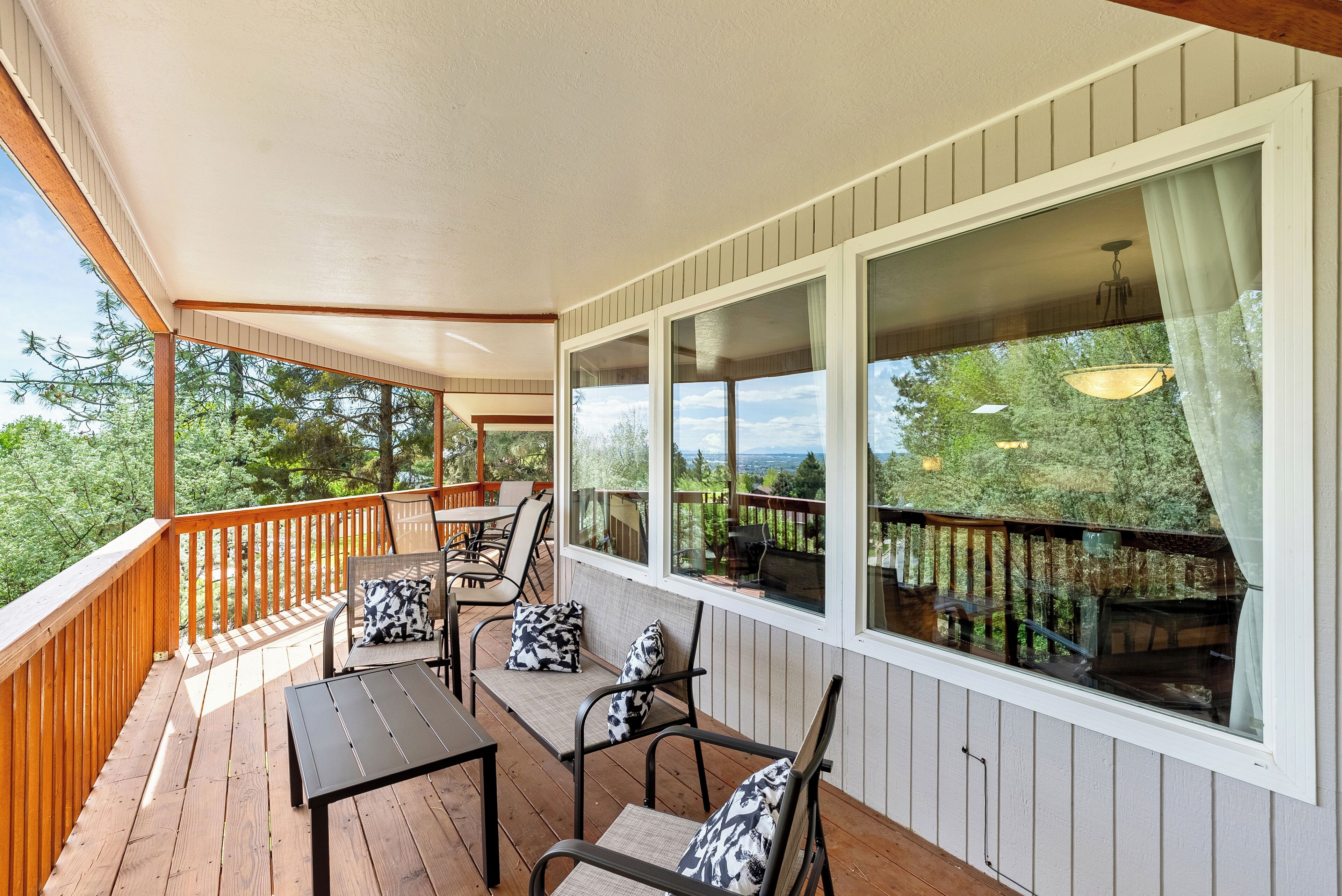 Eine überdachte Terrasse im Freien mit Holzboden, stahlrahmenartigen Stühlen mit gemusterten Kissen und einem Tisch. Große Fenster mit Vorhängen bieten Blick auf die umliegende Grünfläche.