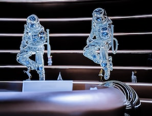 Deux sculptures en verre représentant des astronautes, sur un fond sombre agrémenté de lignes horizontales.