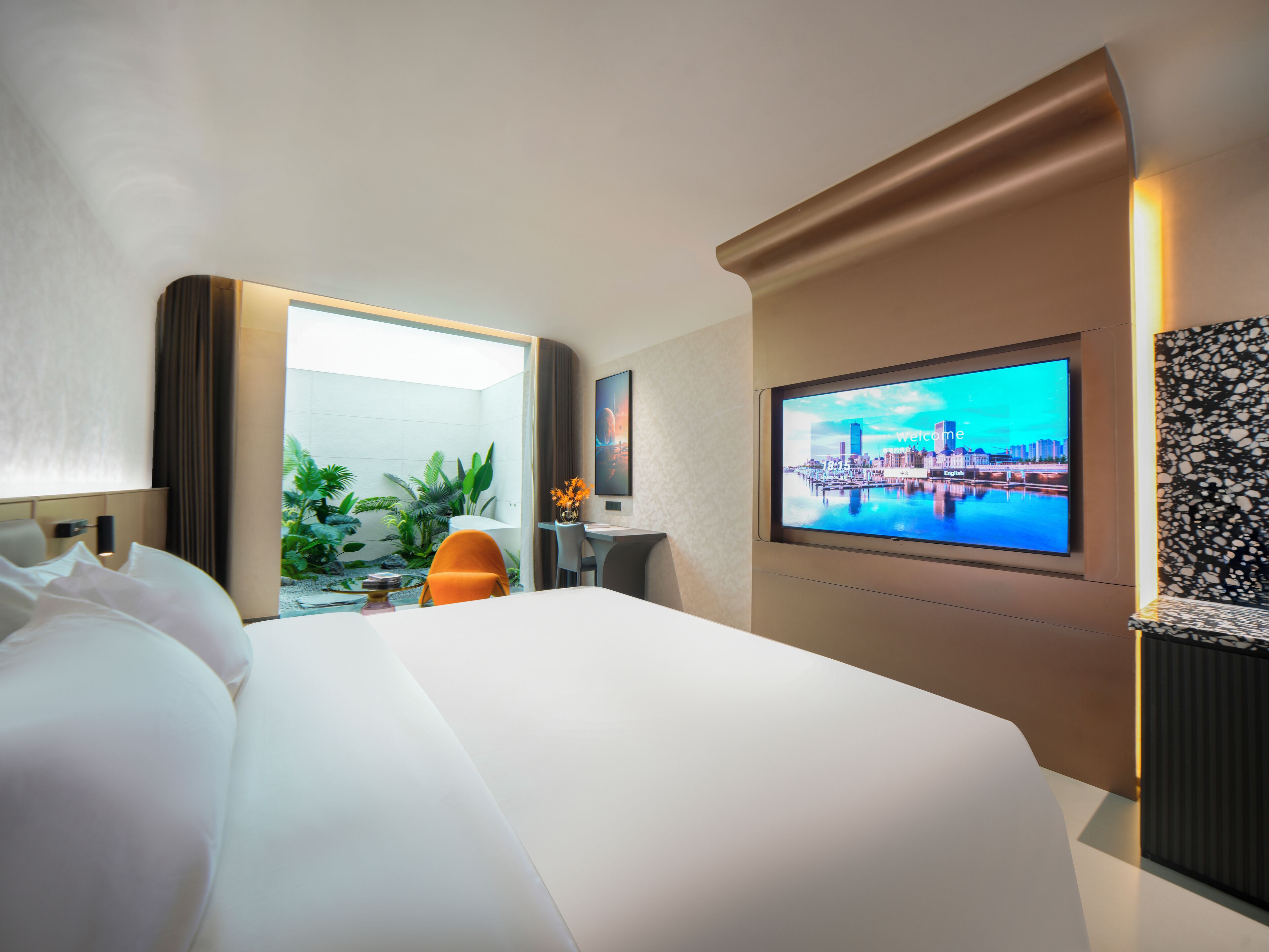 Habitación de hotel con una cama grande, un escritorio con silla, un televisor mostrando una vista urbana y un jardín visible a través de una puerta abierta.