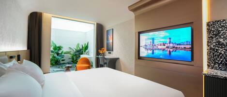 Habitación de hotel con una cama grande, un escritorio con silla, un televisor mostrando una vista urbana y un jardín visible a través de una puerta abierta.