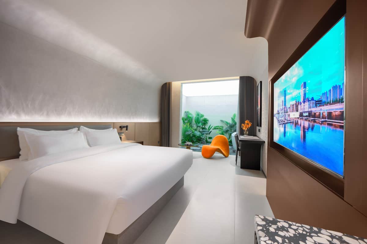 A modern hotel room with a large bed, a TV displaying a cityscape, a small orange chair, and a view of greenery through a window.