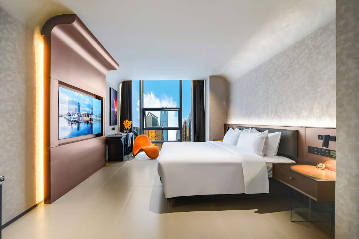 A modern hotel room with a large bed, a desk, a chair, and a view of the city through the window.