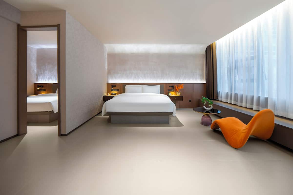 A modern hotel room with a large bed, bedside tables, a small seating area, and a window with sheer curtains.
