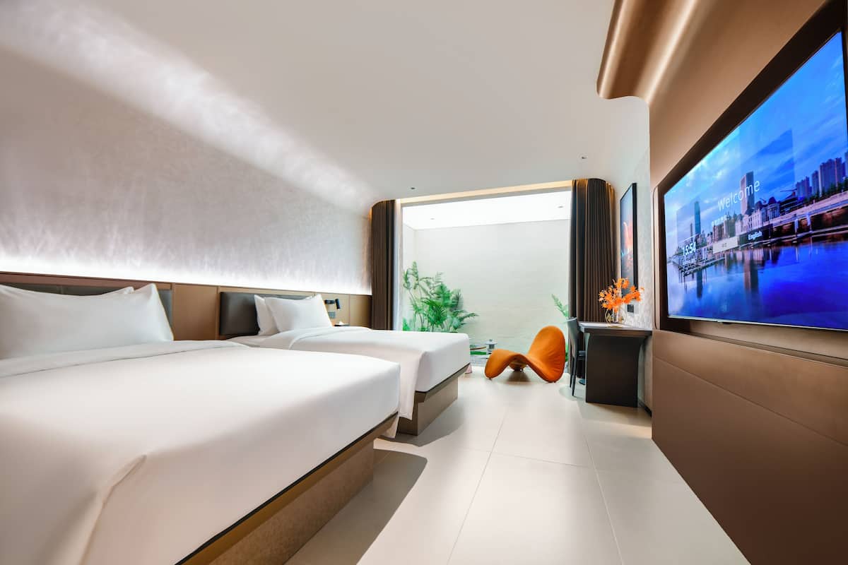 A modern hotel room with two beds, a TV, and a view of the cityscape.