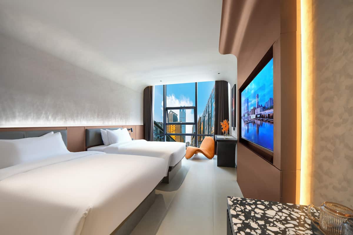 A modern hotel room with two beds, a large TV screen, and a view of the cityscape.