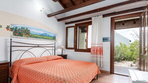 Un dormitorio con cama, mesita de noche, lámpara, un cuadro, ventana con cortinas y vistas al exterior.