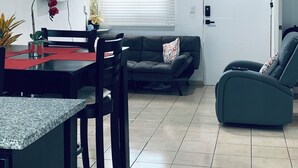 Una sala de estar moderna con comedor, que incluye un sofá, un sillón reclinable, un televisor montado en la pared y una ventana con persianas.