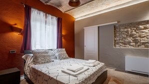 A bedroom with a bed, a nightstand, a wall-mounted light, a window with curtains, and a stone wall feature. - Apartment 'Il Rifugio Illuminato - Morellino' with Shared Garden, Wi-Fi and Air Conditioning (Semproniano)