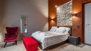 A bedroom with a bed, a red chair, a red ottoman, a stone wall, and a white door. - Apartment 'Il Rifugio Illuminato - Brunello' with Shared Garden, Wi-Fi and Air Conditioning (Semproniano)