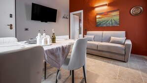 A modern living room with a sofa, a dining table with a chair, a flat-screen TV mounted on the wall, and a painting of a vineyard. - Apartment 'Il Rifugio Illuminato - Brunello' with Shared Garden, Wi-Fi and Air Conditioning (Semproniano)