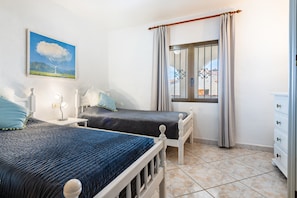 A bedroom with two beds, a window with curtains, a bedside table with a lamp, and a painting on the wall.