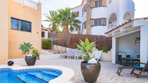 Una piscina con sillas de descanso y plantas en macetas, un área de estar techada y un edificio con balcones.