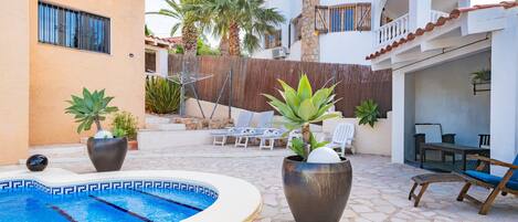 A swimming pool with lounge chairs and potted plants, a covered seating area, and a building with balconies.