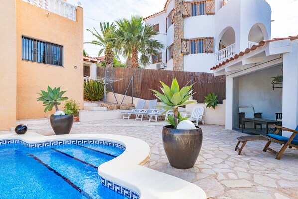 Una piscina con sillas de descanso y plantas en macetas, un área de estar techada y un edificio con balcones.