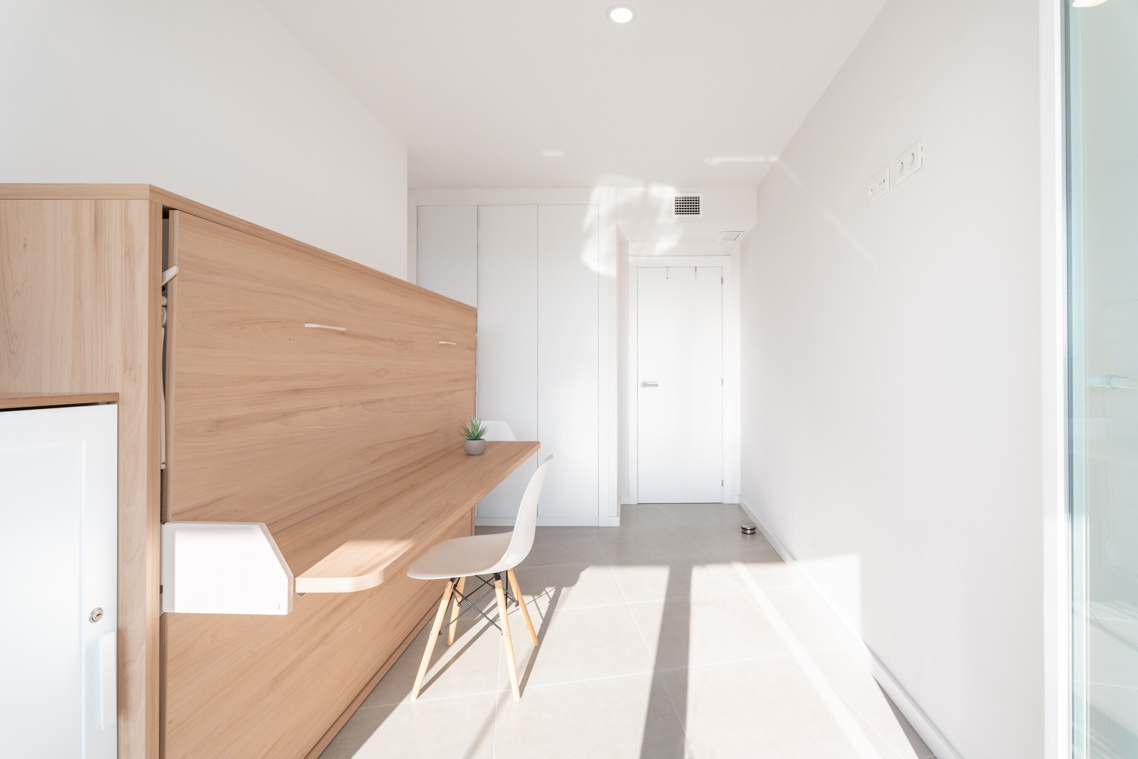 Una habitación moderna con un escritorio de madera, una silla blanca, una planta pequeña y una puerta blanca.