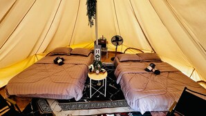 A tent with two beds, each with a pillow and a rolled-up towel. There is a small table with a plant and a picture frame.