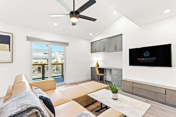 A modern living room with a sofa, a coffee table, and a ceiling fan. There is a kitchen area with cabinets and a TV displaying 'FunQuest Vacation Rentals'.