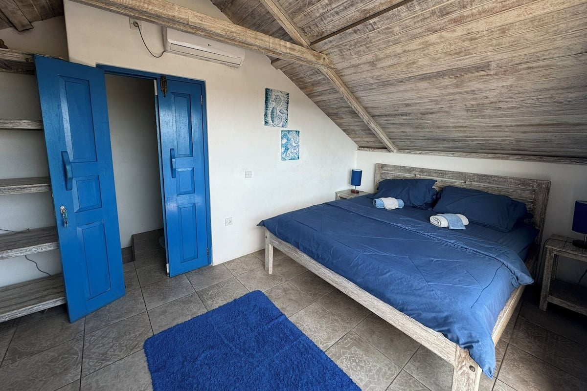 A bedroom with a wooden ceiling, a blue door, a bed with blue bedding, and a small bedside table with a lamp.
