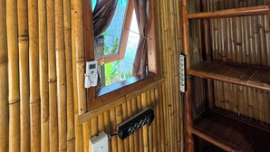 A room with bamboo walls, a window with curtains, a wall-mounted air conditioner, and a shelf with a trash bin. - Bali Bio Villas (Ungasan)