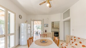 A room with a dining table, chairs, a sofa, a ceiling fan, a refrigerator, and a view of the outside.