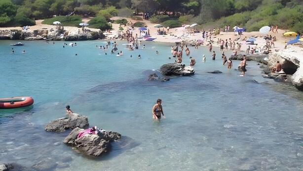 Une plage avec une eau claire, des rochers, et des gens qui se baignent et prennent des bains de soleil.