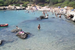 Une plage avec une eau claire, des rochers, et des gens qui se baignent et prennent des bains de soleil.