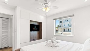 A hotel room with a bed, a ceiling fan, a TV, and a window with a view of buildings.