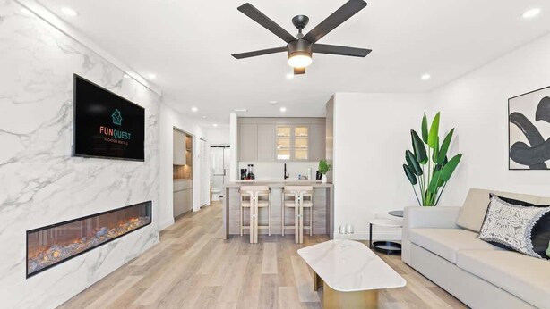 A modern living room with a flat-screen TV, a ceiling fan, a fireplace, a kitchen with bar stools, and a sofa with a decorative pillow.