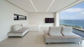 A modern, minimalist bedroom with a large window overlooking the sea, a flat-screen TV mounted on the wall, and a comfortable seating area with a sofa and two armchairs. - Geoje Lighthouse Pension (Geoje)