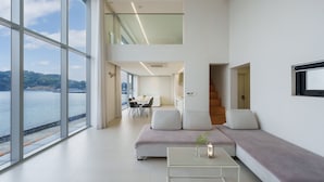 A modern living room with a large window overlooking a body of water, a dining area, and a staircase leading to an upper level. - Geoje Lighthouse Pension (Geoje)