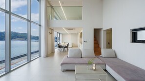 A modern living room with a large window overlooking a body of water, a dining area, and a staircase leading to an upper level. - Geoje Lighthouse Pension (Geoje)
