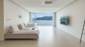 A modern living room with a sofa, a flat-screen TV, and a large window overlooking a body of water. - Geoje Lighthouse Pension (Geoje)