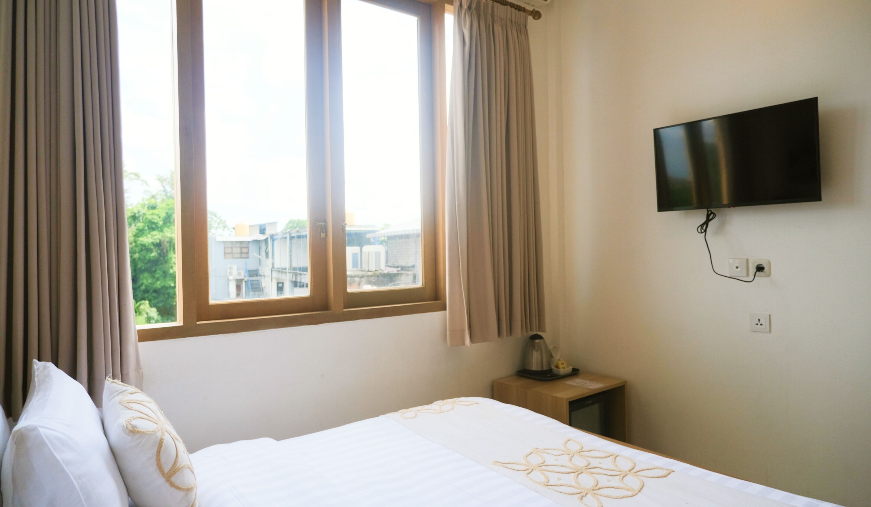 A hotel room with a bed, a TV mounted on the wall, a window with curtains, and a small table with a kettle.