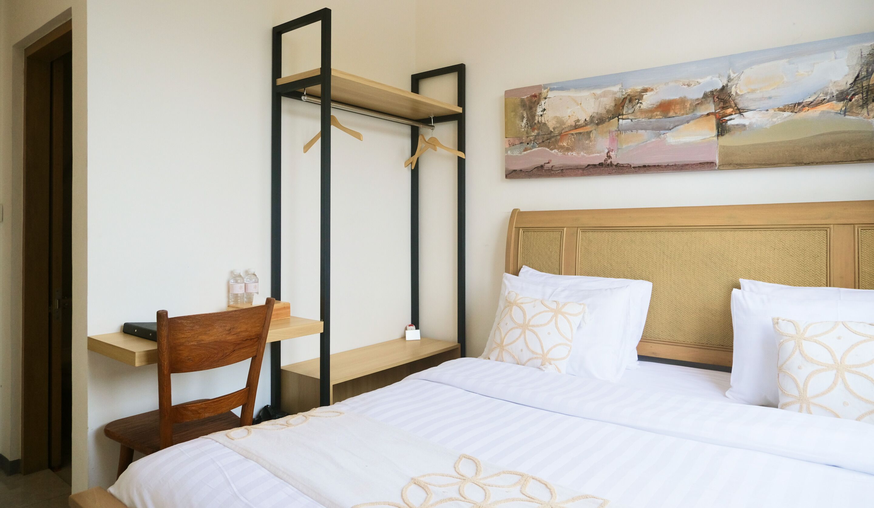 A hotel room with a bed, a wooden chair, a desk with a chair, a hanging shelf, and a painting on the wall.