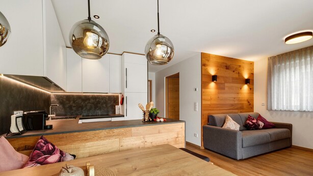 Une cuisine moderne avec un plan de travail en bois, une table à manger avec un set de table, et un canapé dans un coin salon agrémenté d’un mur d’accent en bois et d’une fenêtre avec des rideaux.