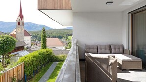 Un balcon avec un canapé en osier, une vue sur une église et une chaîne de montagnes en arrière-plan.