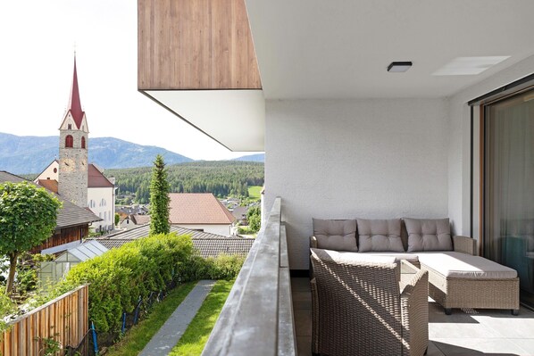 Un balcon avec un canapé en osier, une vue sur une église et une chaîne de montagnes en arrière-plan.